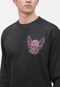 Gros plan d'une personne portant un sweat-shirt noir avec un emblème ailé violet et le mot "DIESEL" sur la poitrine.