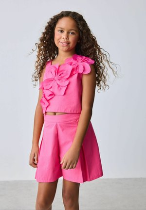 Girl with long curly hair wearing a bright pink sleeveless top with large fabric flowers and matching pleated shorts.