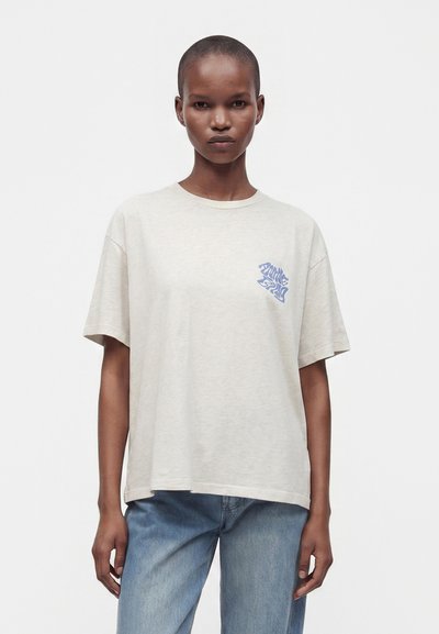 Femme aux cheveux courts portant un t-shirt ample beige clair avec un motif bleu sur la poitrine et un jean bleu clair, debout devant un fond blanc uni.