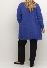 Langer, offen gestalteter Cardigan in lebhaftem Blau, aus strukturiertem Strickstoff. Mit geripptem Saum und seitlichen Taschen. Kombiniert mit schwarzen Hosen und flachen Schuhen.