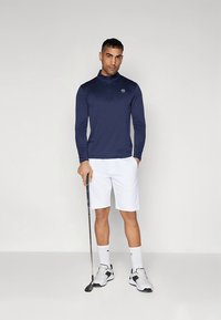 Tengerészkék hosszú ujjú galléros póló félcipzárral, fehér rövidnadrággal párosítva. Kiegészítőként egy golfütő. Cipők fehér-feketék.