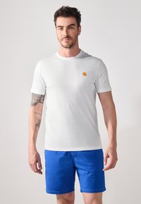 White short-sleeve t-shirt made of cotton, featuring a small orange pattern on the chest, paired with blue shorts. Simple, clean design.