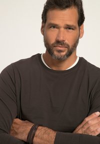 Maglietta scura a maniche lunghe con scollatura rotonda e colletto interno bianco a contrasto. Il modello ha una barba e indossa braccialetti di pelle marrone.