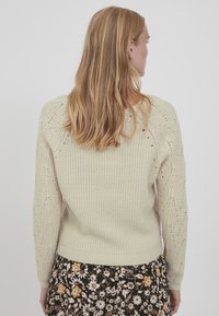Striktrøje i lys beige med et tekstureret mønster og ribbede kanter; har blomstermønstrede detaljer på ærmerne. Kombineret med en nederdel med blomsterpattern.