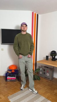 Zöld polár pulóver, világosszürke nadrág és szürke tornacipő. Lila sapka. A szobában színes csíkok, egy TV és egy fából készült asztal dekorációval található.