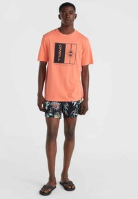 Camiseta gráfica color coral con detalles en negro, combinada con pantalones cortos de baño negros con estampado floral. El modelo lleva sandalias negras, mostrando un atuendo casual de verano.