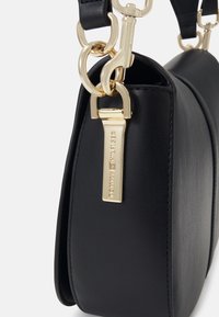 Sac à main en cuir noir vu de côté avec une quincaillerie dorée et une étiquette rectangulaire gravée attachée à une sangle d'épaule noire.