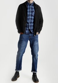 Czarna zamszowa kurtka na niebieskiej koszuli w kratę, zestawiona z niebieskimi dżinsami z podwiniętymi mankietami i czarnymi botkami za kostkę. Prosty i funkcjonalny design.