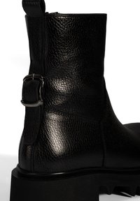 Bottine en cuir noir avec une finition texturée, dotée d'un détail de boucle sur le côté et d'une semelle épaisse et texturée.