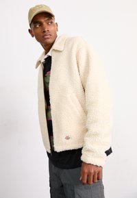 Giacca in pile beige e soffice con colletto, chiusura con cerniera frontale e polsini a costine, indossata sopra una maglietta nera e pantaloni cargo color oliva.