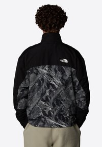 Svart och mönstrad jacka med en texturerad design på baksidan, med hög krage och logodetalj på övre vänstra sidan.