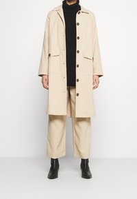 Person, der einen beigen langen Mantel mit Knöpfen, beigefarbene weite Hosen, einen schwarzen Rollkragenpullover und schwarze Stiefeletten vor einem weißen Hintergrund trägt.