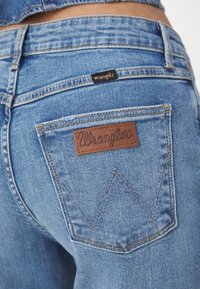 Izbliza prikaz stražnjeg džepa plavih traperica s smešnim kožnim Wrangler logom i ušivenim detaljem "W" na džepu.