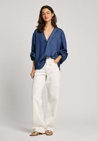 Tunika di jeans blu scuro con scollatura a V e maniche lunghe arrotolate, abbinata a pantaloni larghi bianchi e sandali metallici con cinturini.