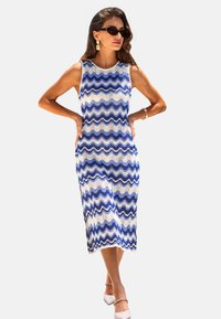 Vestido sin mangas de patrón en zigzag azul y blanco, de longitud hasta la rodilla, con silueta ajustada, fabricado en tela ligera. Lleva unos tacones blancos.