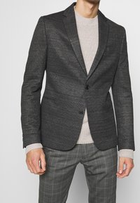 Schwarzer Blazer mit strukturierter Oberfläche, taillierter Passform, Reverskragen und einem einzelnen Knopfverschluss, kombiniert mit einem leicht gestrickten Pullover und karierter Hose.