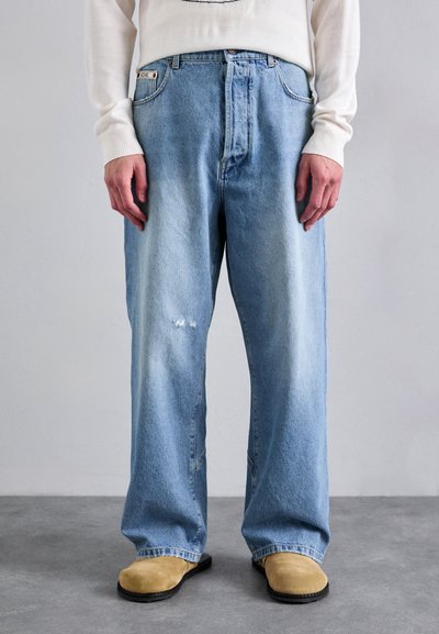 Vaaleansiniset denim-farkut rento malli, joissa on haalistunut rakenne ja pieni kulunut alue. Yhdistettynä ruskeisiin puukengiin, seisten harmaalla lattialla.
