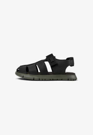 Black sandal with an open design, featuring elastic Velcro strap, rubber sole, and ventilation cutouts. Textured upper and flexible fit.