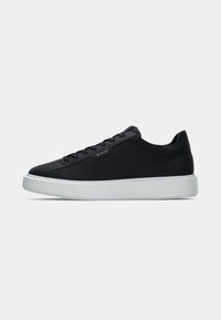 Zwarte leren sneaker met een ronde neus, witte rubberen zool, getextureerde accenten en zes oogjes voor veters. Gouden logo-detail aan de zijkant.