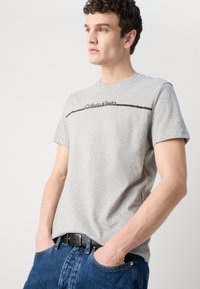 Gray cotton T-shirt with short sleeves, featuring a black Calvin Klein logo and stripe across the chest, paired with blue denim jeans.