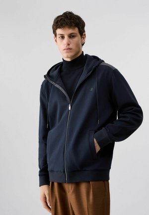 Jeune homme portant un sweat à capuche zippé bleu marine sur un col roulé noir et un pantalon marron, debout devant un fond clair uni.
