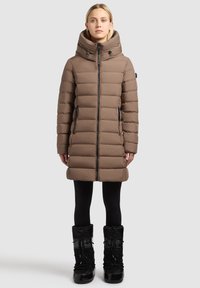 Brown puffer coat with a high collar, vertical quilted design, two side zippers, and a front zip closure. Worn over black leggings and boots.