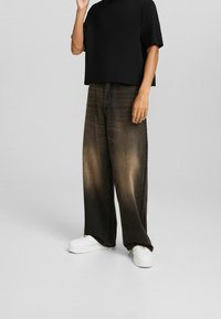 Svart croppad t-shirt kombinerad med lösa, blekbruna jeans. Vita sneakers och en framåtvänd handgest fulländar outfiten.