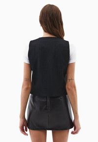 Sort skråstribet cropped vest over en hvid t-shirt, kombineret med en højhælet sort lædernederdel i mini. Glatte teksturer og tætsiddende design.