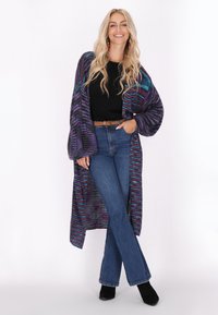 Lång, öppen cardigan i lila, teal och svart stickad, bärs över en svart topp och högmidjade blåjeans, med svarta ankelskor.
