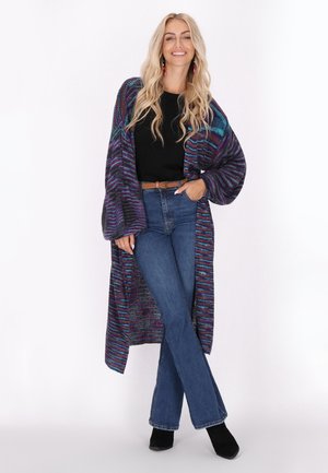 Cardigan lungo aperto in maglia viola, teal e nero, indossato sopra una maglietta nera e jeans blu a vita alta, con stivali neri alla caviglia.