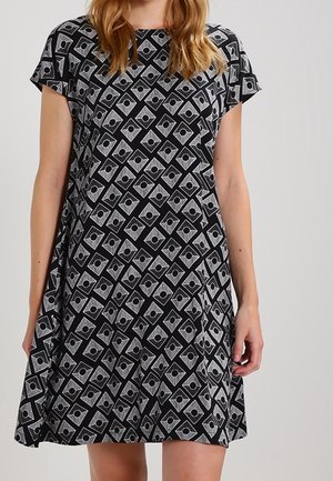 Kobieta ubrana w sukienkę o kroju literą A z krótkim rękawem, ozdobioną czarno-szarym geometrycznym wzorem diamentowym z centralnymi czarnymi okręgami.