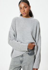 Hellgrauer, gestrickter Pullover mit lockerem Schnitt, gerippter Textur, Rundhalsausschnitt und überschnittenen Schultern. Kombiniert mit Jeans aus Denim mit Nieten.