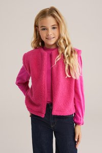 Chlpatá ružová bezrukávová vesta s jemnou textúrou, nosená cez dlhý rukáv ružového topu. Tmavomodré džínsy dotvárajú celý outfit.