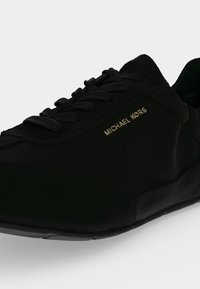 Μαύρο αθλητικό παπούτσι Michael Kors με μαύρα κορδόνια και χρυσά γράμματα λογότυπου στο πλάι, εμφανιζόμενο σε λευκό φόντο.