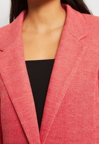 Blazer à motif chevron rouge avec revers à encoches, présentant une silhouette ajustée. Haut noir en dessous, affichant une texture lisse.