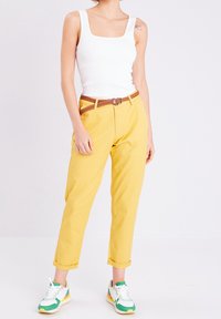 Pantalon en coton jaune avec une ceinture en cuir tressée marron, associé à un débardeur blanc à côtes et des baskets vertes, blanches et oranges.