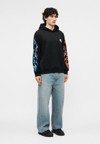 Tânăr îmbrăcat cu un hanorac negru cu flăcări albastre pe mâneca stângă, flăcări roșii pe mâneca dreaptă, blugi deschiși la culoare și pantofi negri, într-o poziție neutră.