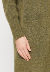 Stickad grön klänning med långa ärmar, som har en subtil textur och en sidoficka, med en hand vilande på tyget nära fållen.