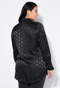 Zwarte blouse met een gestructureerd honingraatpatroon, lange mouwen en een kraag. De stof lijkt glad en licht glanzend.