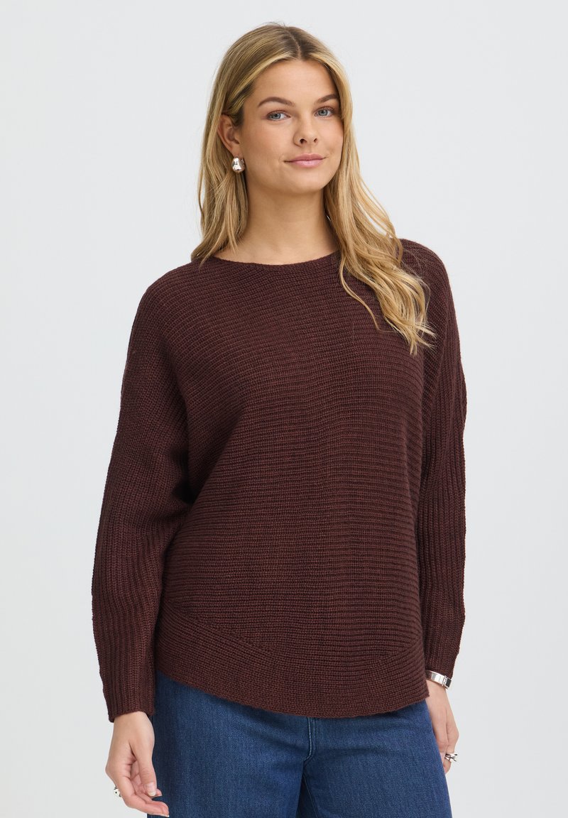 Femme aux longs cheveux blonds porte un pull en maille marron ample et un jean bleu, debout devant un fond clair et uni, regardant devant elle.