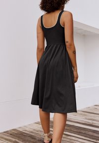 Fekete ruha testhezálló felső résszel és bővülő szoknyával. Széles pántokkal és sima, könnyű anyagból készült. Jelentős varrás a nyakkivágás mentén.