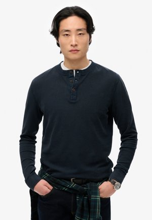 Camisa henley de manga larga en color marino con tejido texturizado, tapeta de tres botones y cuello redondo. Camisa de cuadros oscuros anudada a la cintura.