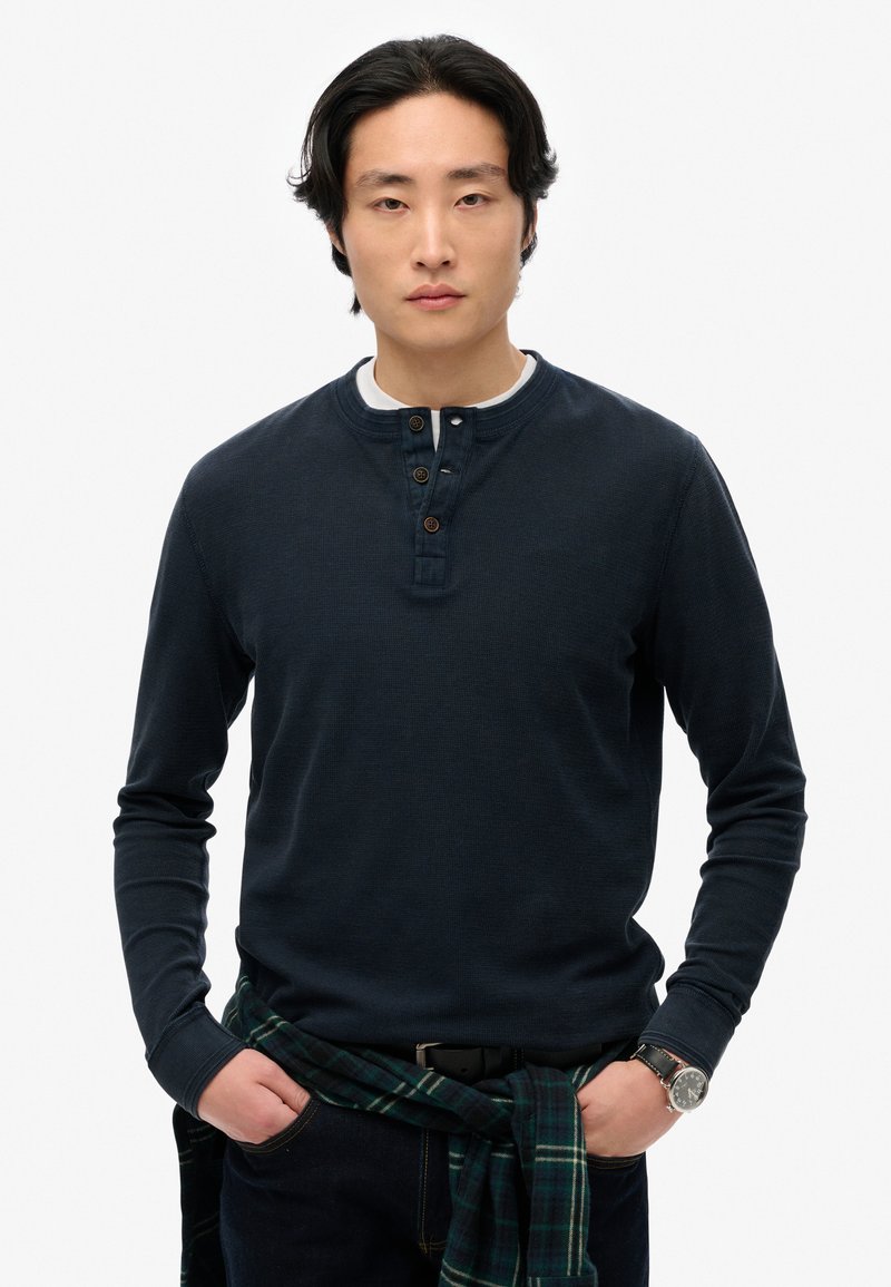 Camicia henley a maniche lunghe blu navy con tessuto testurizzato, fermata con tre bottoni e scollatura a girocollo. Camicia a quadri scura legata in vita.