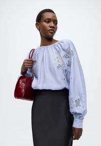 Ανοιχτό μπλε μπλούζα με φουσκωτά μανίκια και λεπτομέρειες από δαντέλα με λουλούδια, συνδυασμένη με μαύρη φούστα. Κρατείται μια μικρή δερμάτινη τσάντα σε μπορντό.