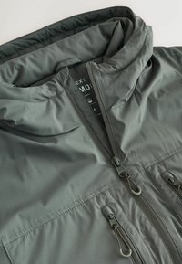 Jacket impermeabilizant gri cu glugă, având un fermoar complet, două buzunare frontale și ajustări cu șnur. Textură netedă și finisaj mat.