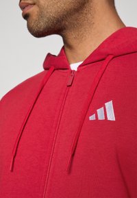 Κόκκινο φούτερ με φερμουάρ, κατασκευασμένο από μαλακό ύφασμα, διαθέτει φερμουάρ στο μπροστινό μέρος, κορδόνια και λευκό κεντημένο λογότυπο στο στήθος.