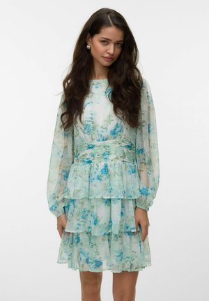 Kvinde med langt mørkt hår iført en lyseblå og grøn blomsterlangærmet lagdelt kjole, stående mod en ensfarvet hvid baggrund.