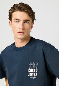 Camiseta de algodón azul marino con cuello redondo. Presenta un estampado gráfico con "CASEY JONES" y tres figuras en el lado izquierdo del pecho.