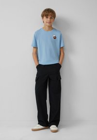 Lichtblauwe katoenen T-shirt met korte mouwen en een patch; gecombineerd met zwarte cargobroek met zakken en platte schoenen met een witte bovenkant.