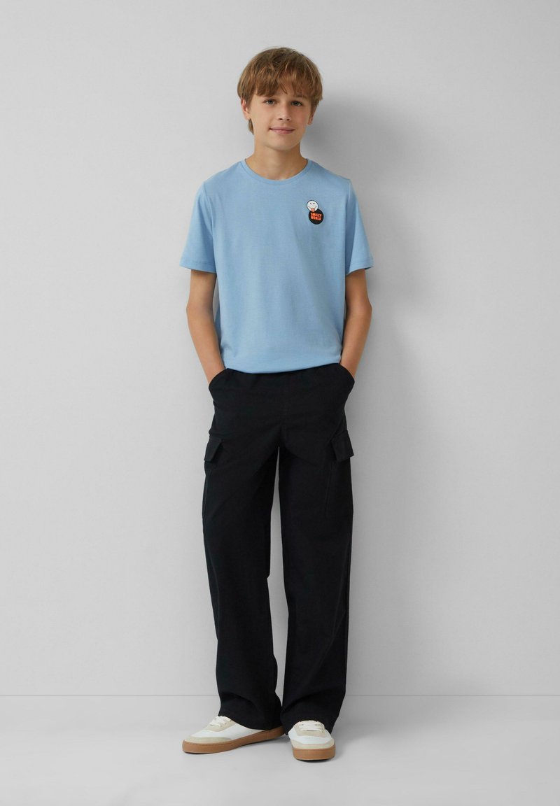 Lichtblauwe katoenen T-shirt met korte mouwen en een patch; gecombineerd met zwarte cargobroek met zakken en platte schoenen met een witte bovenkant.